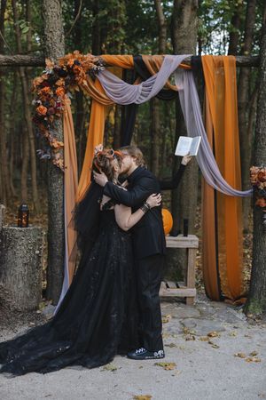 A couple in black gothic wedding attire embrace under an orange and white draped arch in a forest setting.