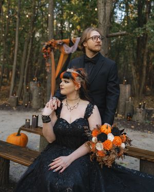 A couple in gothic-inspired wedding attire pose together in a wooded setting with orange and black floral accents.