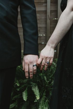 Close-up view of hands wearing multiple dark rings against black clothing and green ferns.
