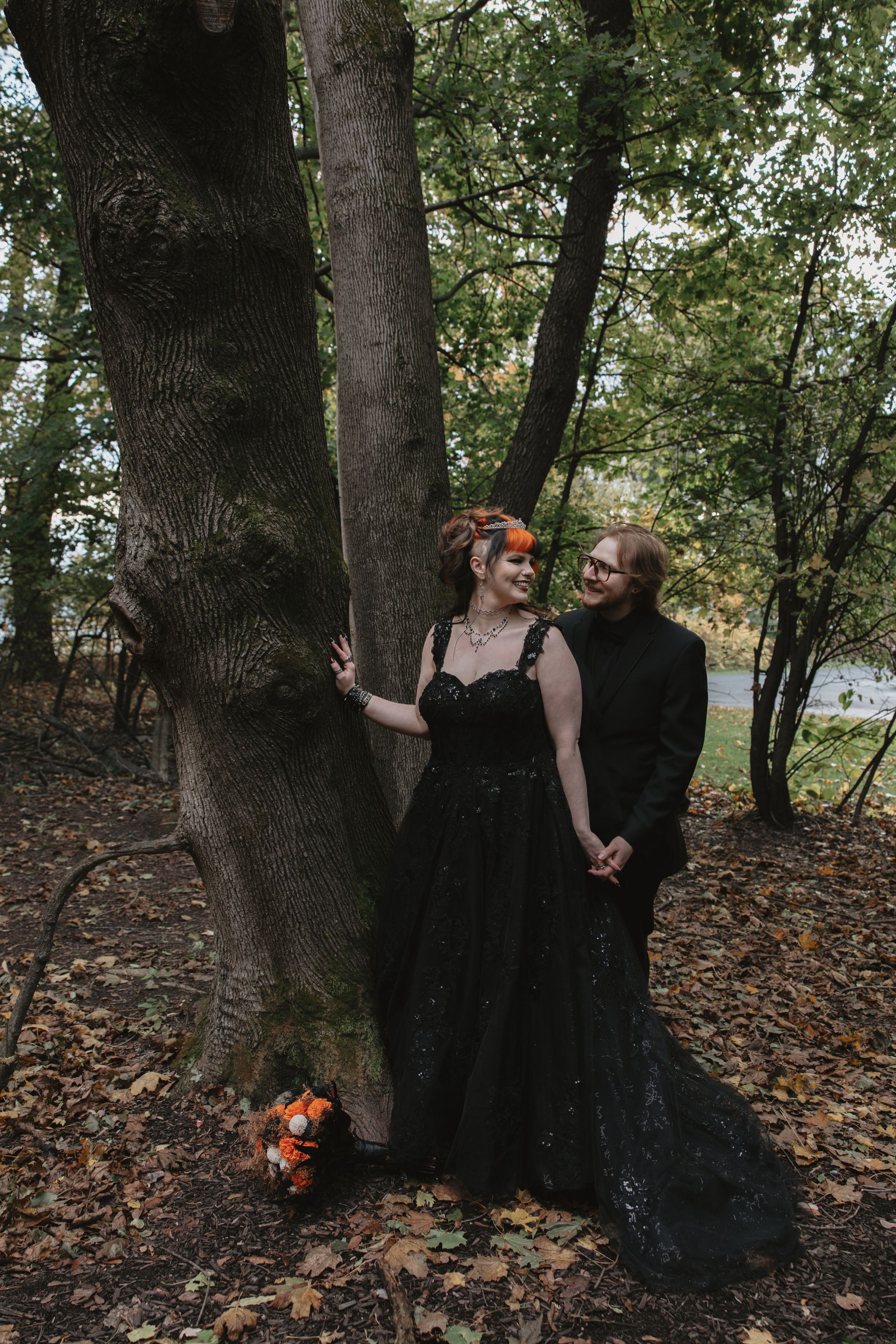 A couple in black outfits poses playfully among autumn trees and fallen leaves.