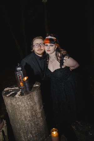 A couple poses with a bottle of Jack Daniels whiskey on a rustic tree stump in a dark outdoor setting with candlelight.