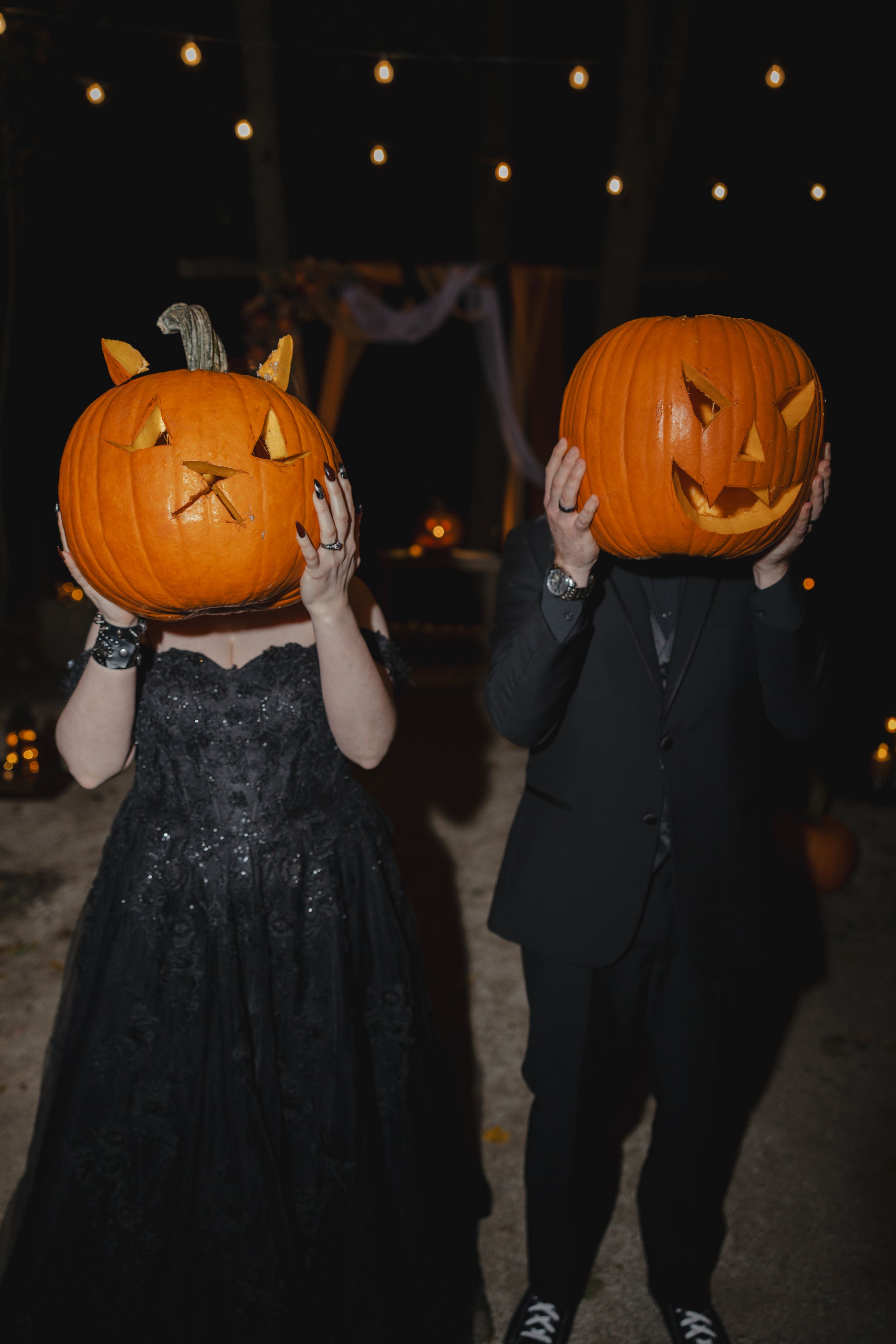 Two people in dark attire hold carved jack-o'-lanterns at night with string lights in the background.