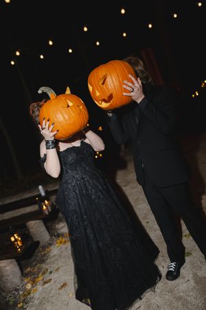 A couple in black attire holding carved pumpkins over their faces at night with string lights in the background.