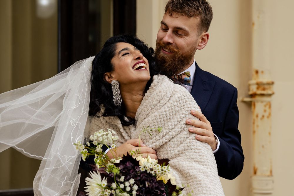 Bride wearing a cozy white sweater laughs joyfully while holding wedding bouquet with purple and white flowers.