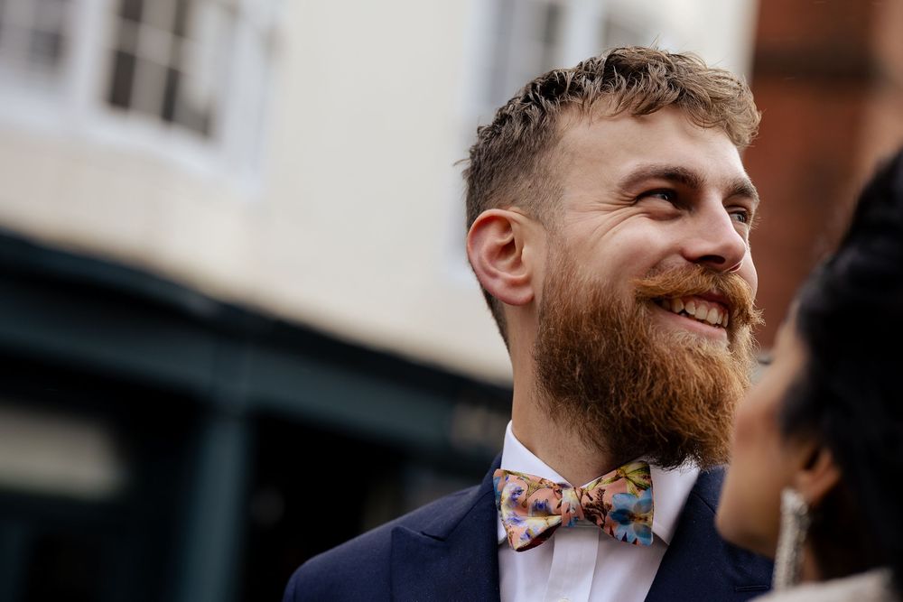 A smiling person with a ginger beard wearing a navy suit and floral bow tie stands outside a building.