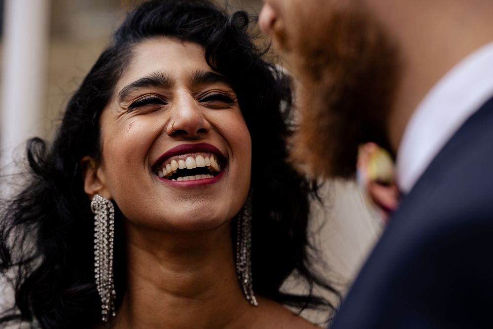 A woman wearing a white dress and dangling earrings smiles joyfully while looking to the side.