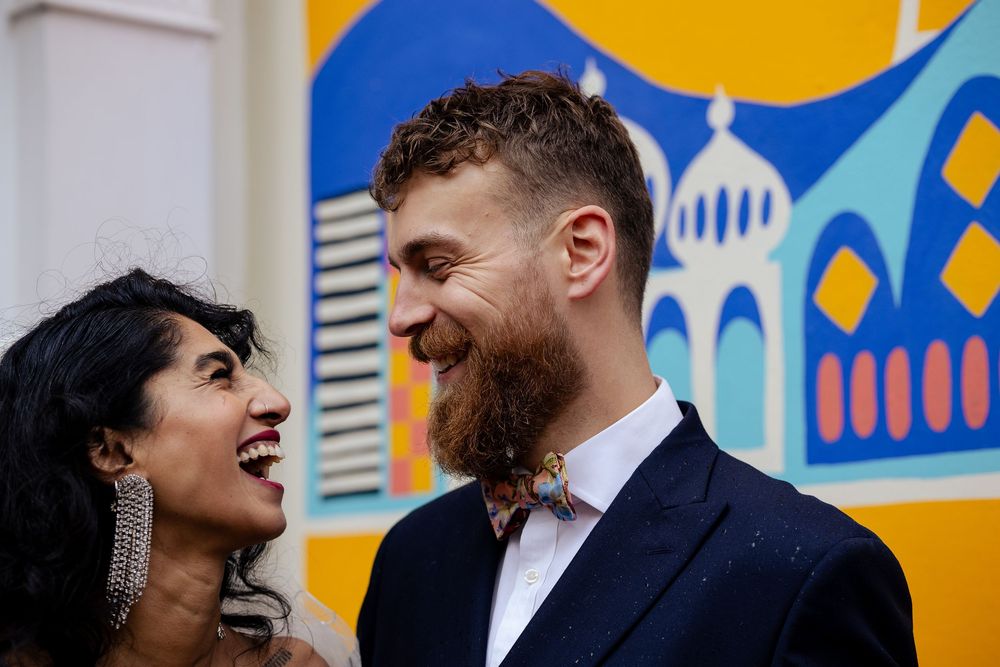 Newlyweds share joyful laughter against a vibrant mural with blue and yellow colors.