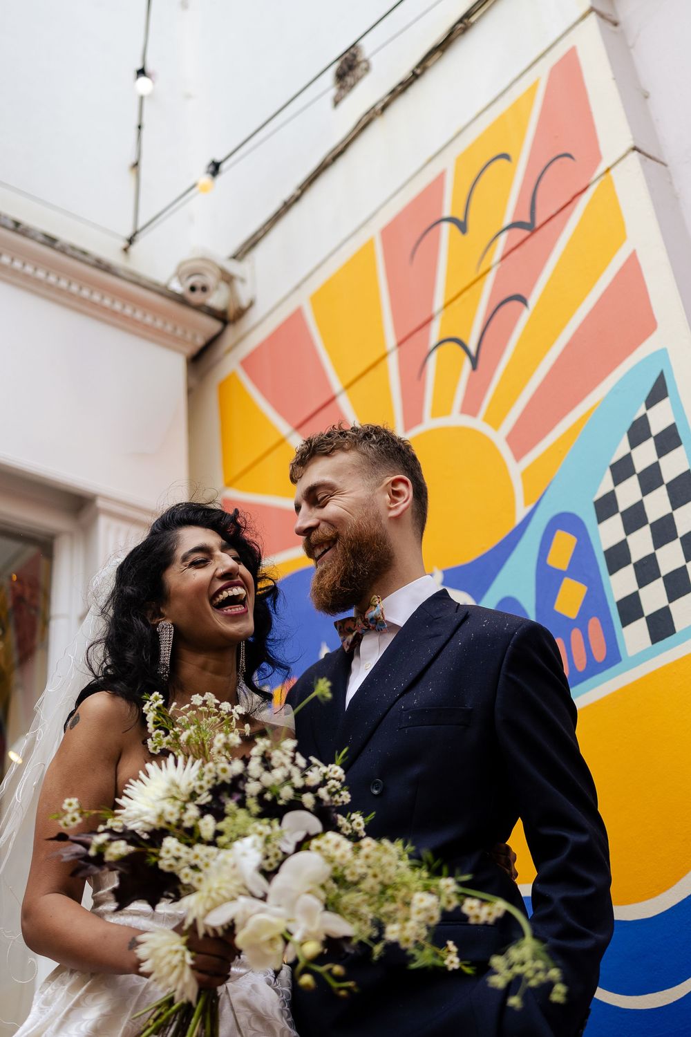 Wedding couple poses with white floral bouquet against colorful art deco style mural.