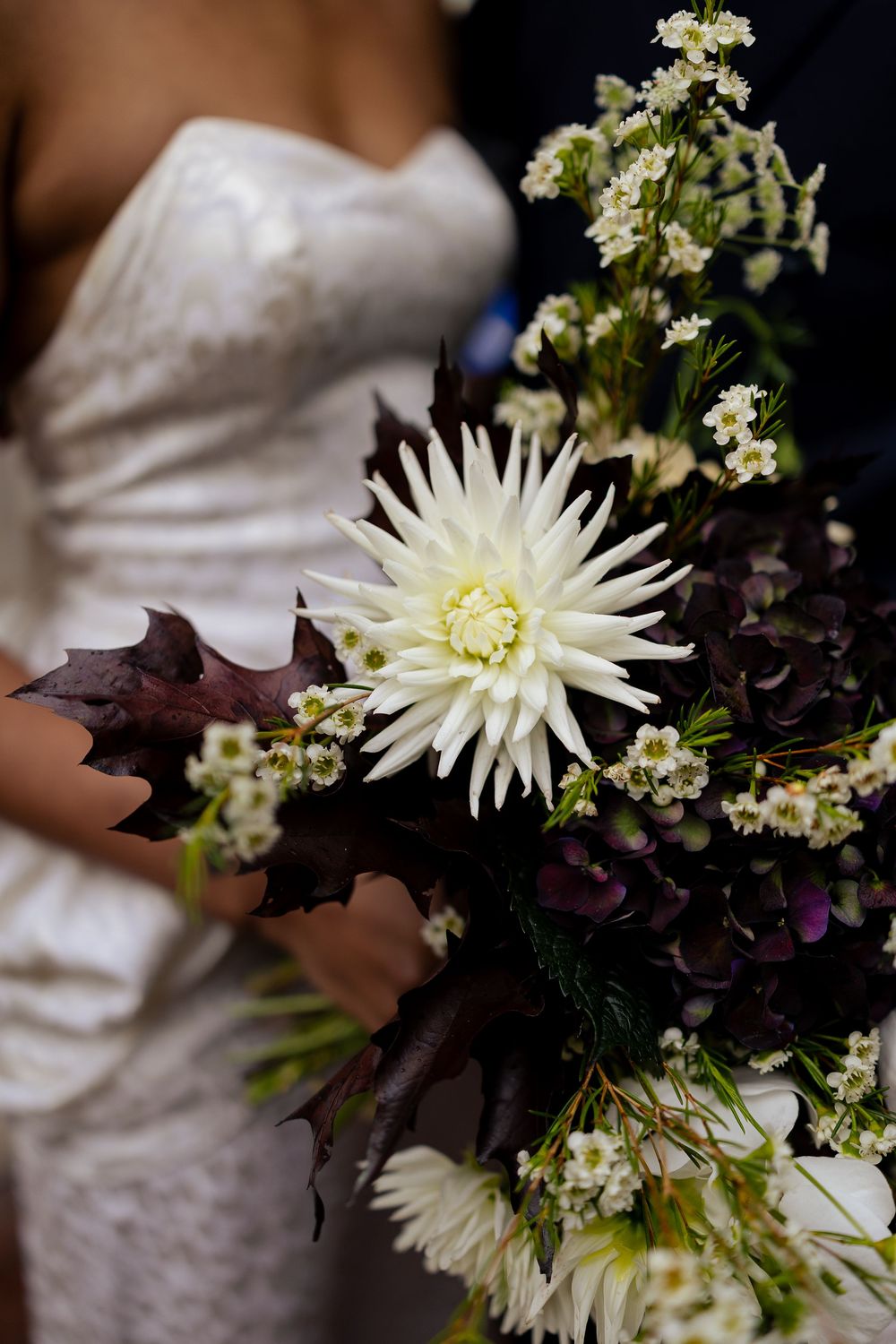 An elegant wedding bouquet with white spider mums, dark purple leaves, and delicate baby's breath against a dark background.