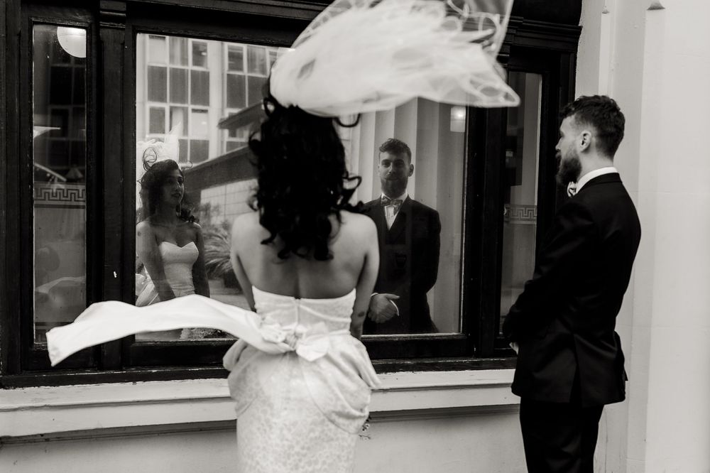 Black and white photo of a bride in a white dress looking through a window while someone stands outside.