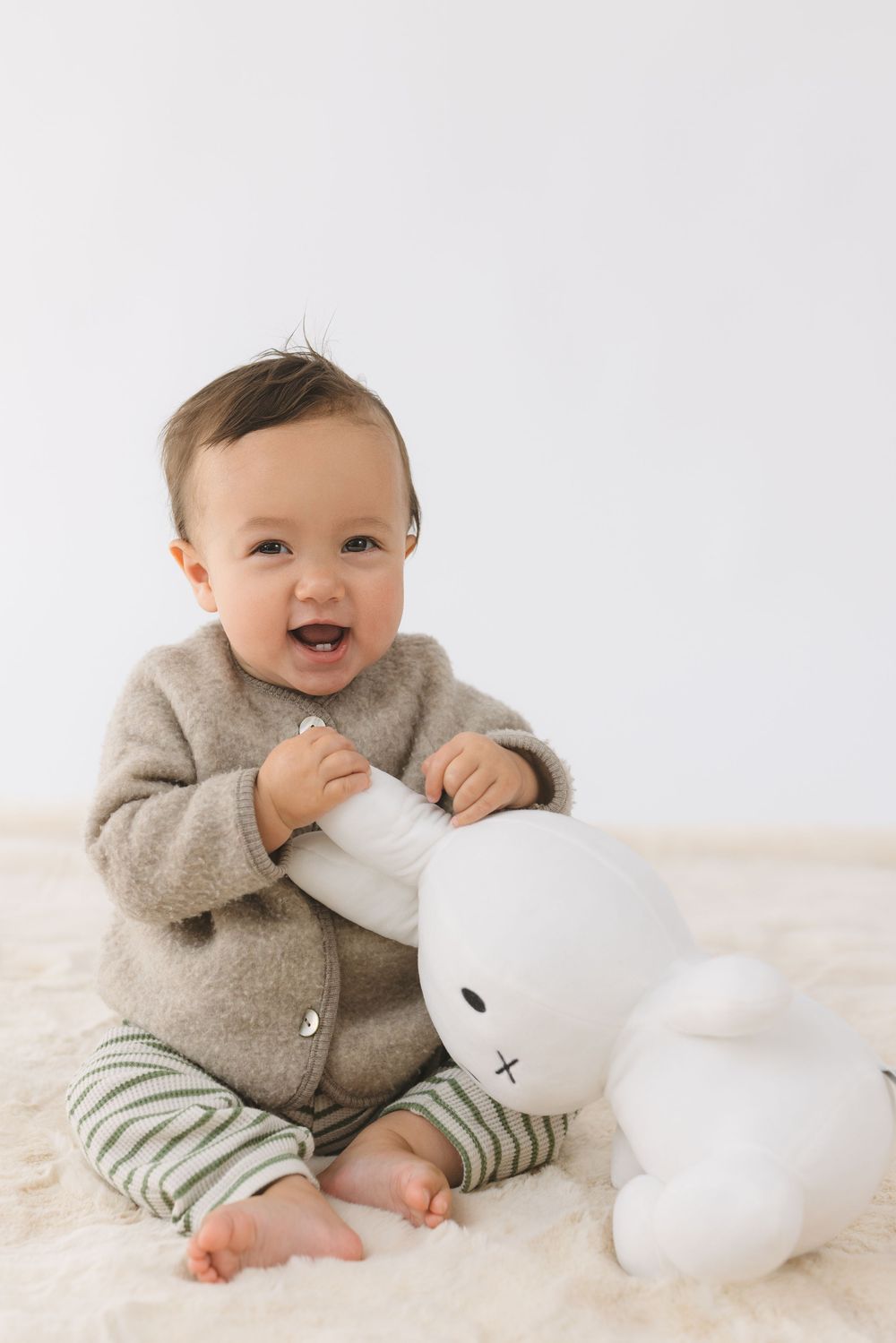 Baby in gray cardigan and striped pants plays with white plush toy on cream blanket.