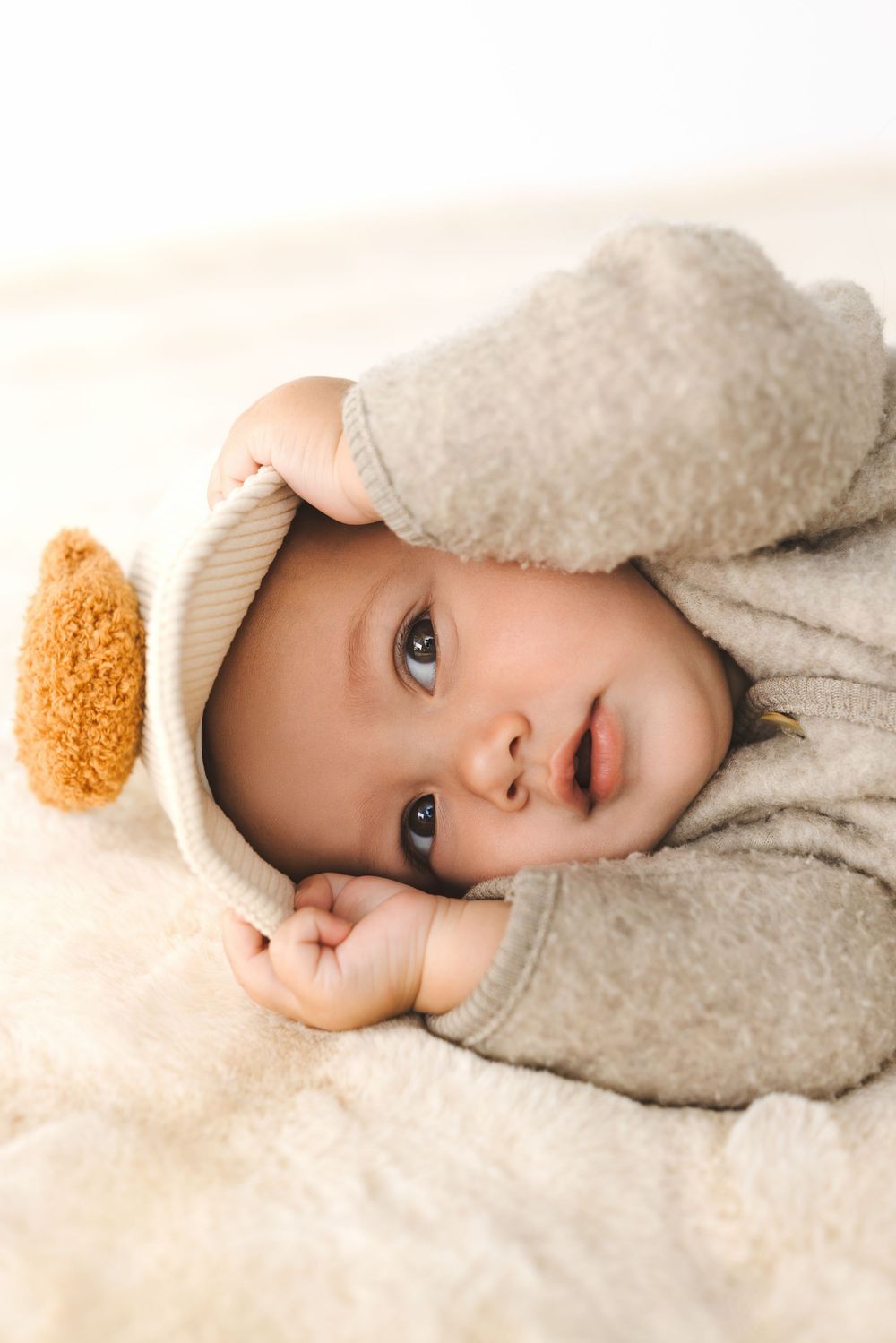 Baby wearing cozy gray hoodie lying on soft white blanket with teddy bear detail.