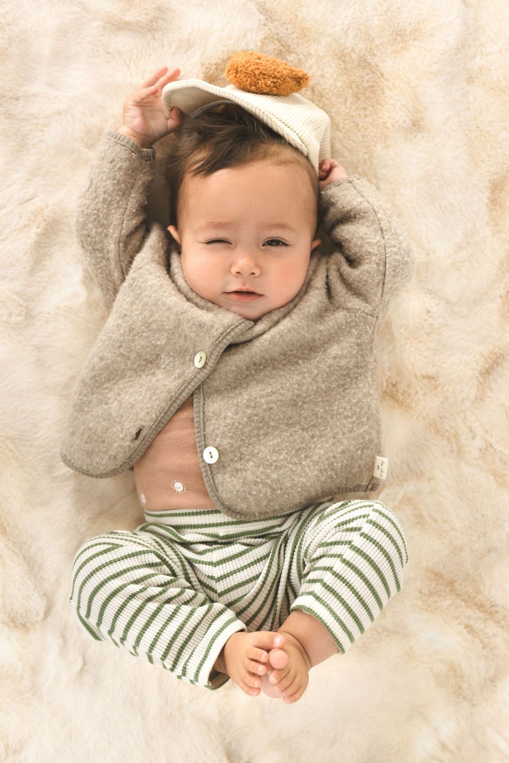Baby wearing gray sweater and striped pants resting on beige blanket in cozy setting.