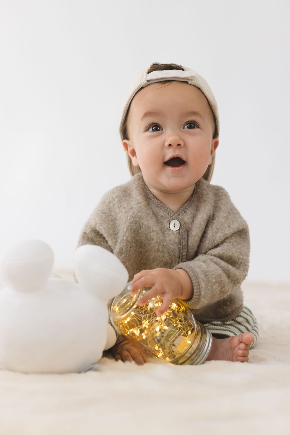 Baby in cozy gray sweater plays with golden Christmas ornament and white decorations on light background.