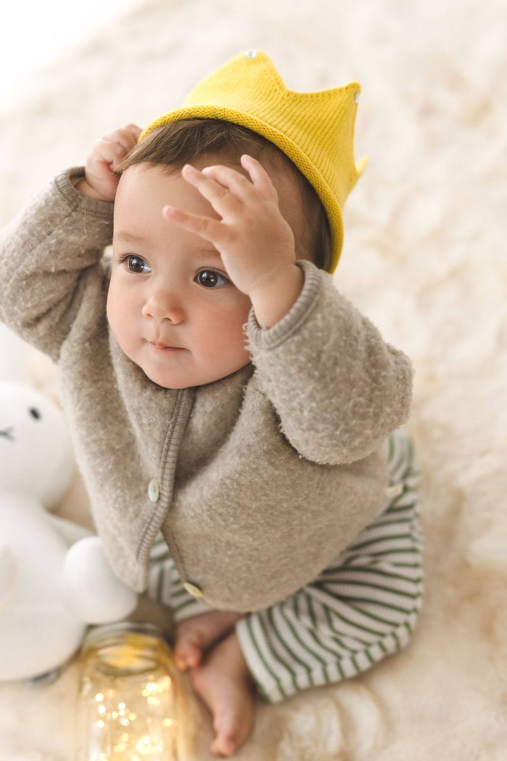 Baby wearing yellow beanie and gray sweater plays with fairy lights on white blanket.