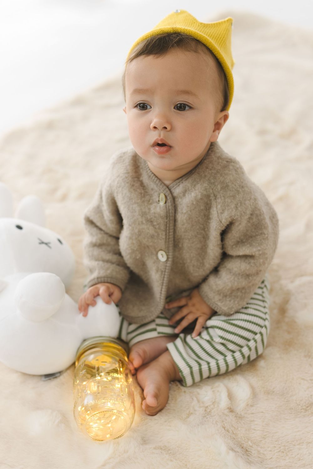 Baby in cozy gray cardigan and striped shirt reaches for glowing light on cream blanket.