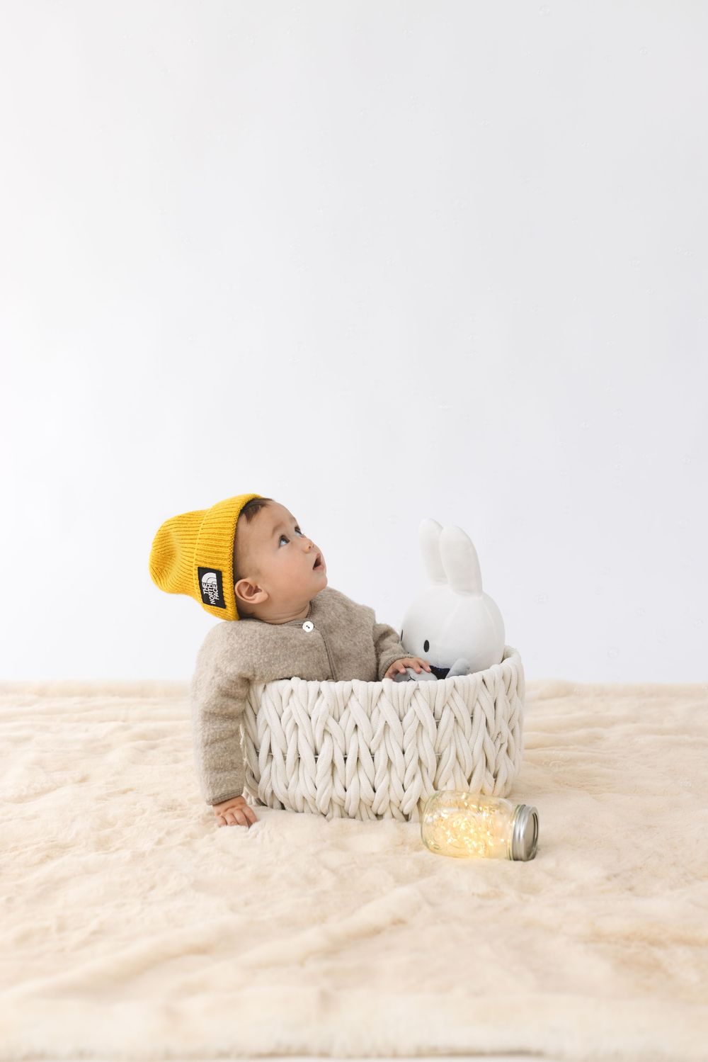 Baby wearing yellow beanie rests in woven basket with white plush bunny and holiday lights.