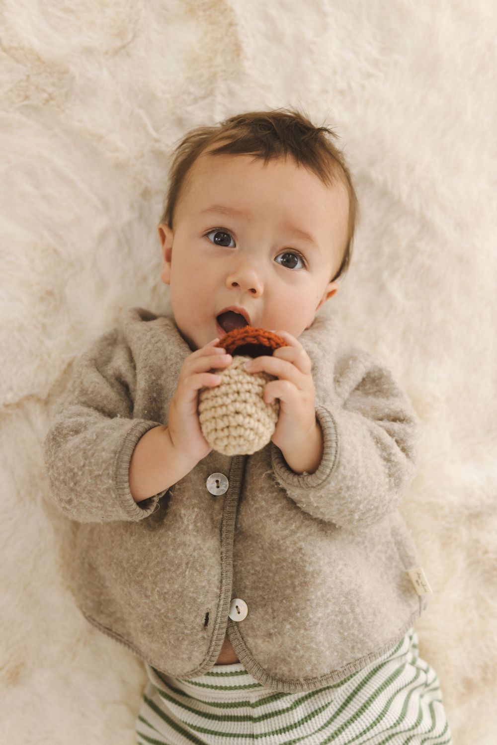 Baby in gray sweater and striped shirt plays with knitted toy on white fuzzy blanket.