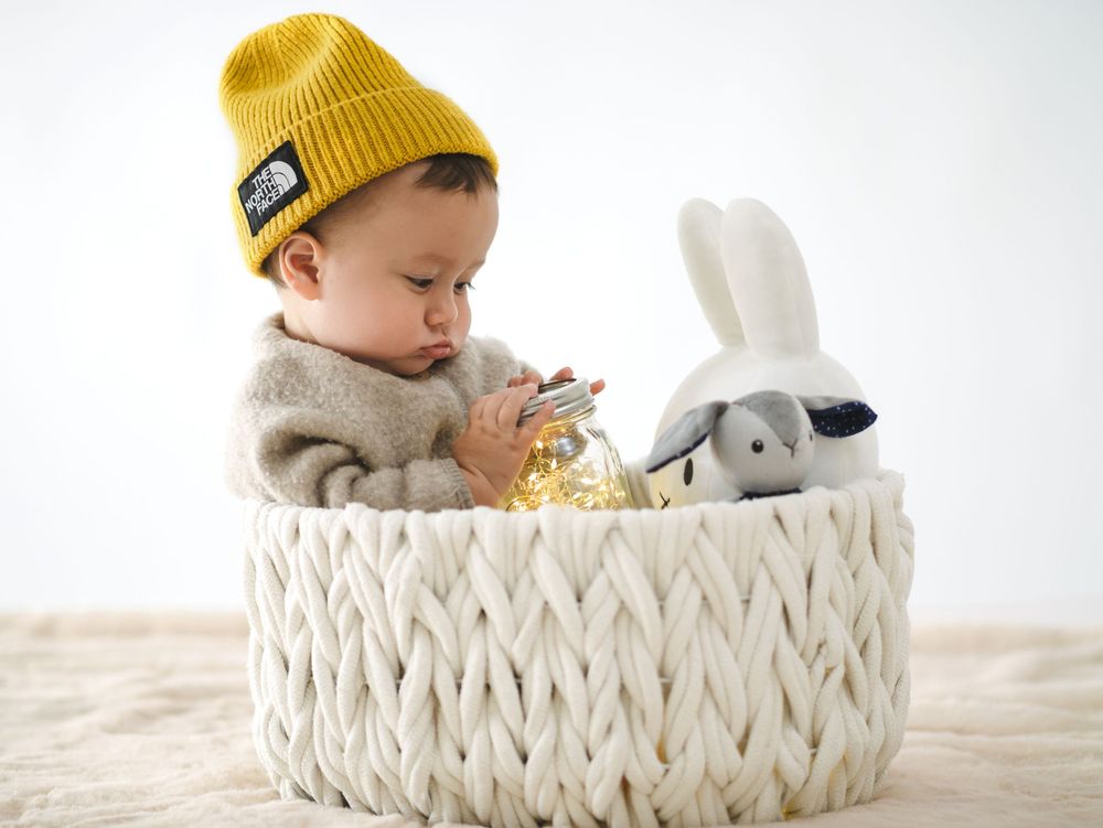 A baby wearing a yellow beanie sits in a white woven basket with a plush bunny toy beside them.