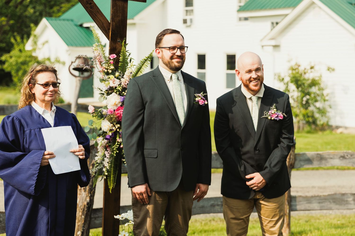 Wedding ceremony takes place under a rustic wooden arch decorated with flowers in a scenic outdoor setting.