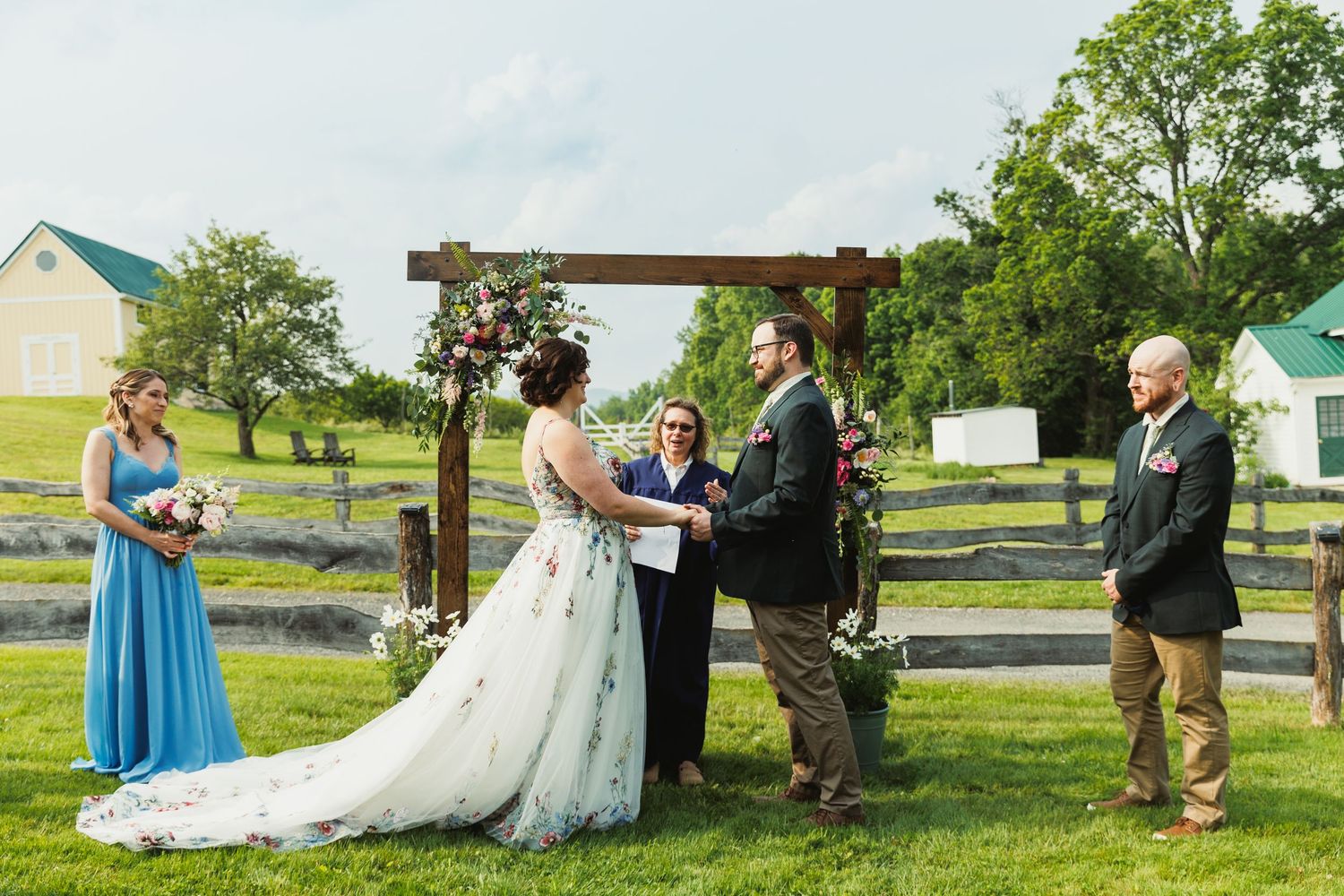 Outdoor wedding ceremony in progress with guests seated on wooden chairs facing a floral-decorated arch.