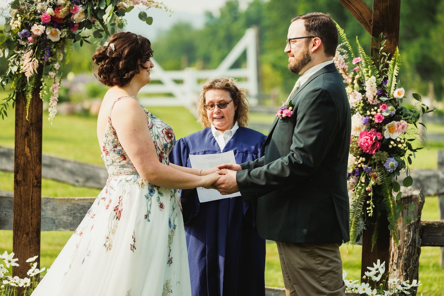 Couple exchanges vows under a wooden pergola adorned with pink and white flowers during outdoor ceremony.
