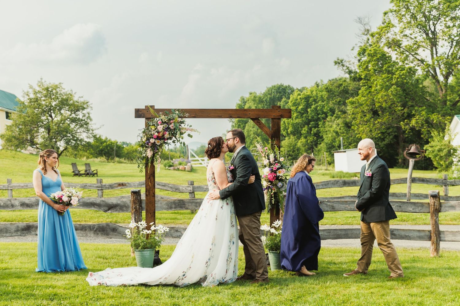Panoramic view of outdoor wedding ceremony with guests seated on lawn near white farmhouse.