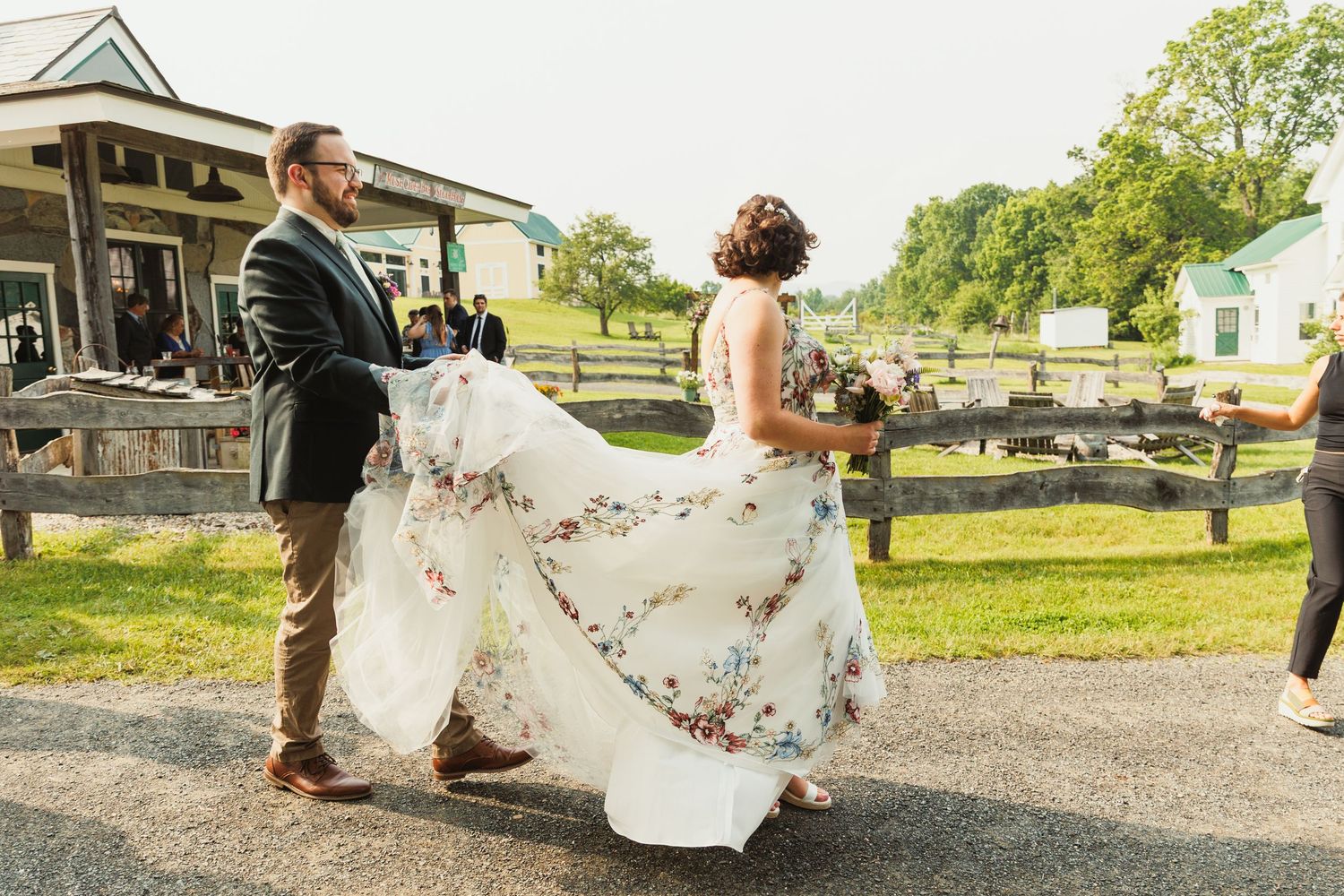 Wedding party poses on gravel path with white farmhouse and green landscape in background.