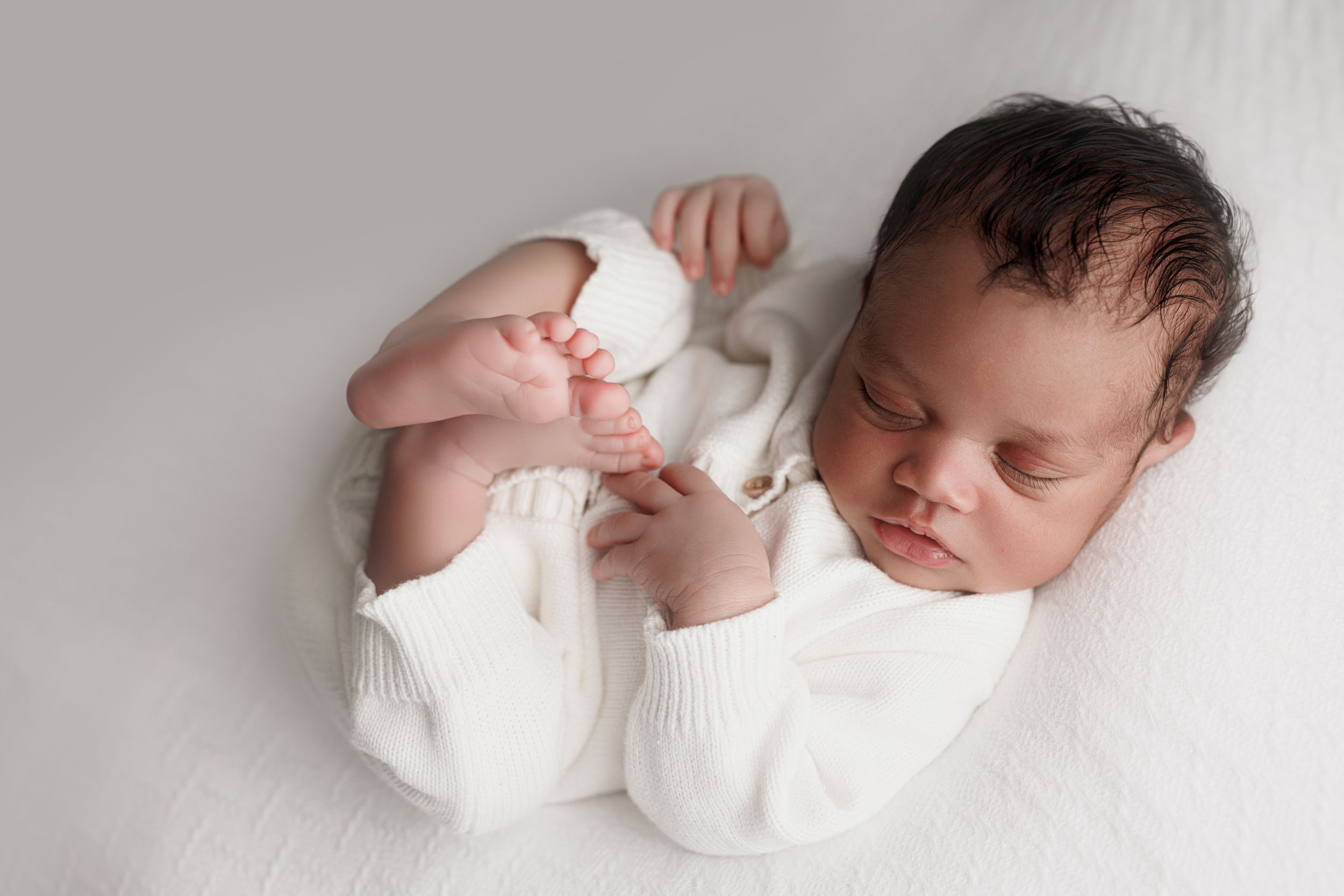 Sleeping newborn baby wearing white onesie lying on soft white blanket in peaceful pose.
