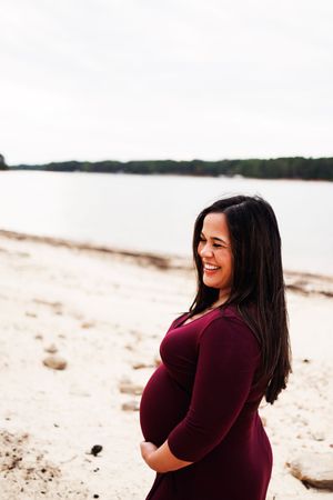 A person in a burgundy sweater smiles while standing on a sandy beach with water and trees in the background.