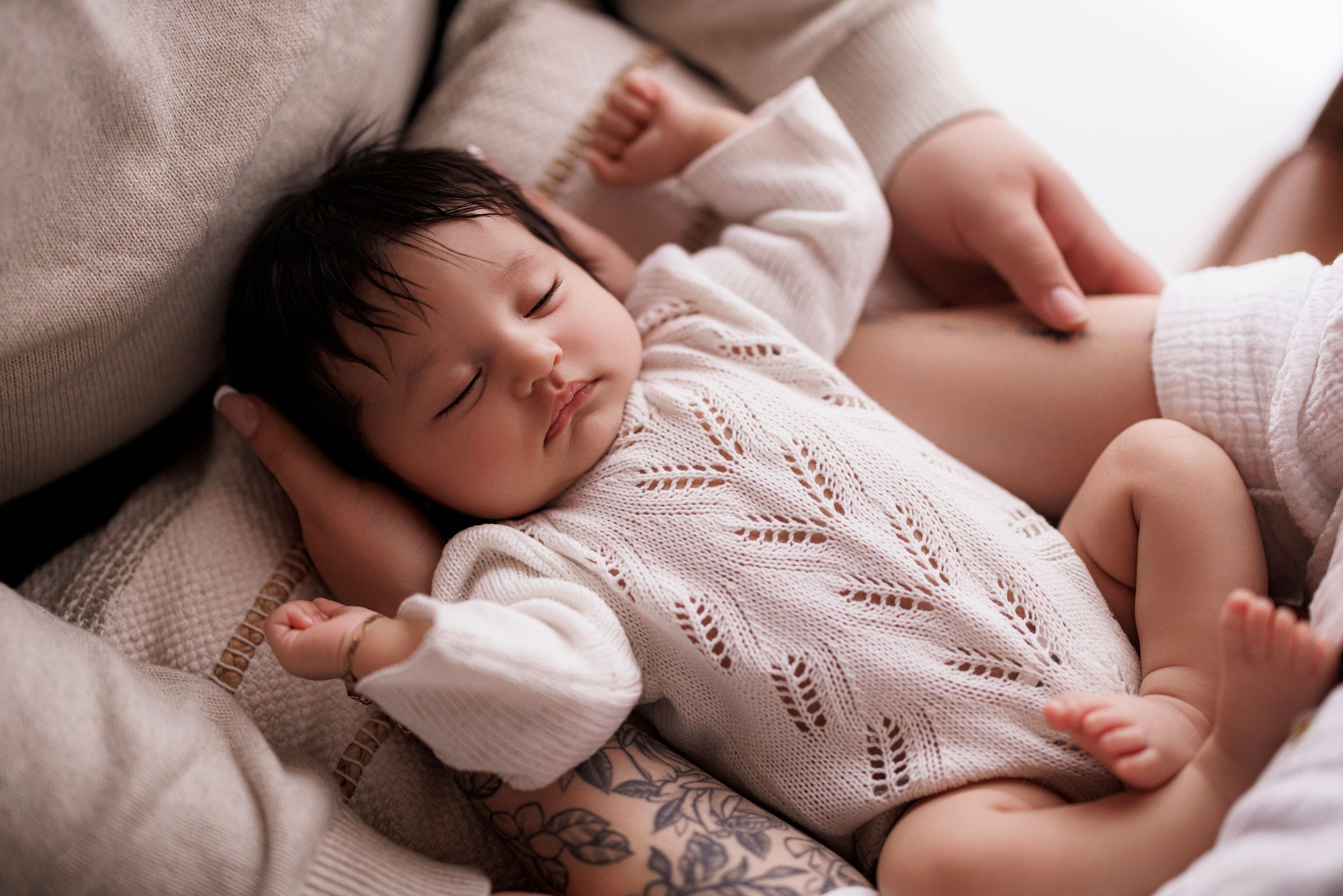 A sleeping baby wearing a white knit sweater rests peacefully on soft bedding.
