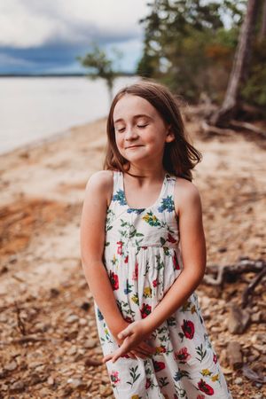 A young girl in a floral dress stands on a rocky beach shoreline during sunset.