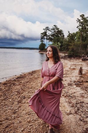 A person in a flowing mauve dress poses on a rocky beach shoreline at sunset.