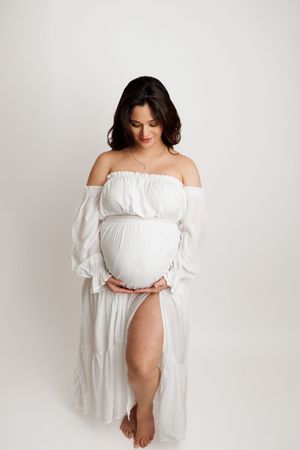 Maternity photo series showing a woman in flowing white off-shoulder dress against light backdrop in elegant poses.
