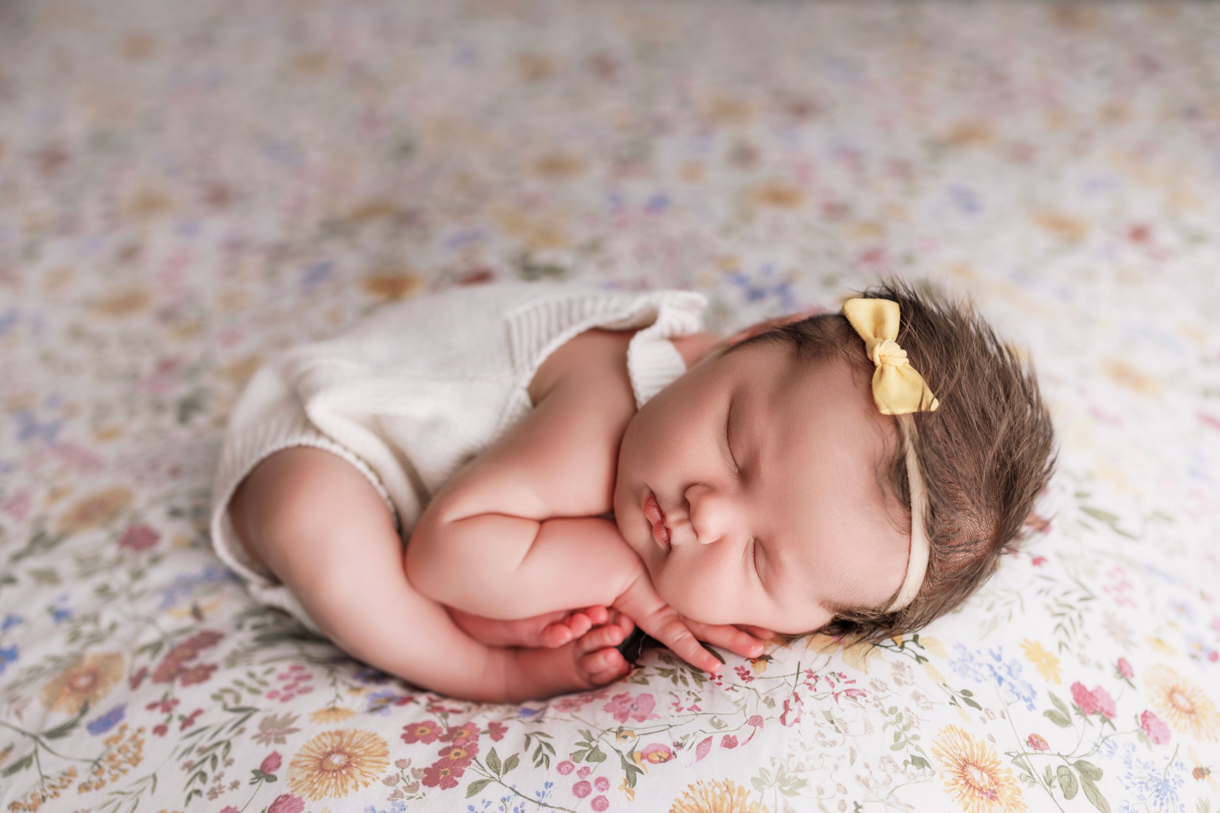 Sleeping newborn in white onesie and yellow bow rests peacefully on floral patterned blanket.