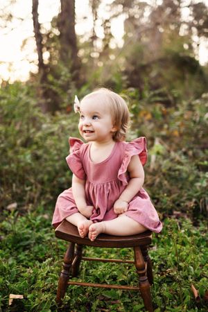 A small child in a pink ruffled dress sits on a wooden stool outdoors surrounded by greenery at sunset.
