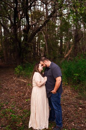 A couple shares an intimate moment together in a wooded forest setting wearing casual attire.