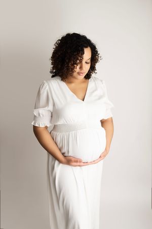 A pregnant person in a white dress poses against a neutral background in a series of maternity photos.