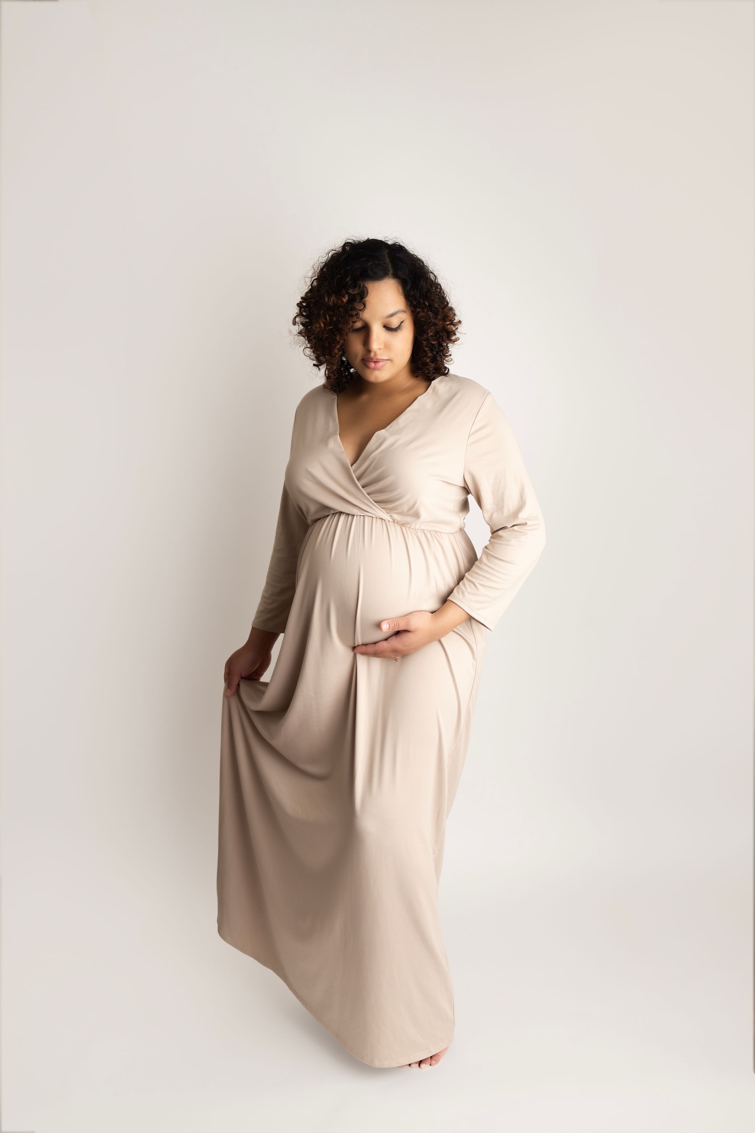 Expectant woman in beige long-sleeved wrap maxi dress poses elegantly against white background during maternity photoshoot.
