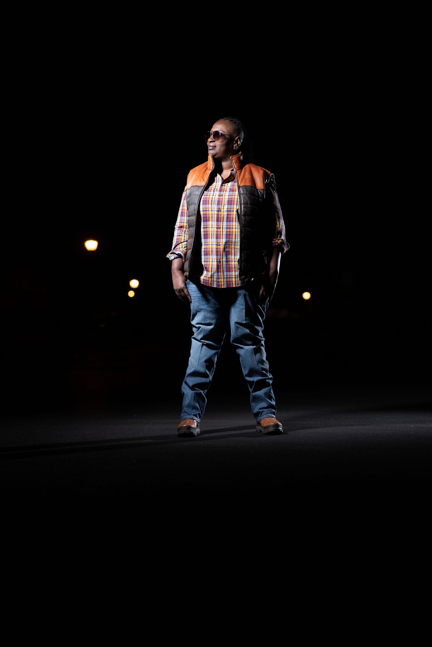 Construction worker in safety vest and plaid shirt stands dramatically lit in dark setting.