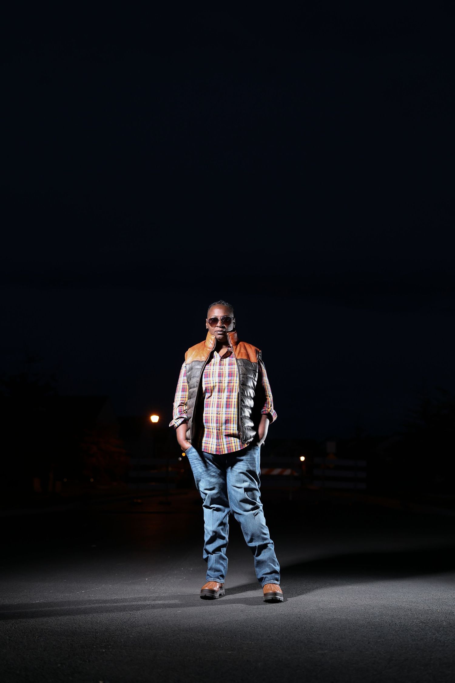 Person in work attire and sunglasses poses under dramatic spotlight on dark background.