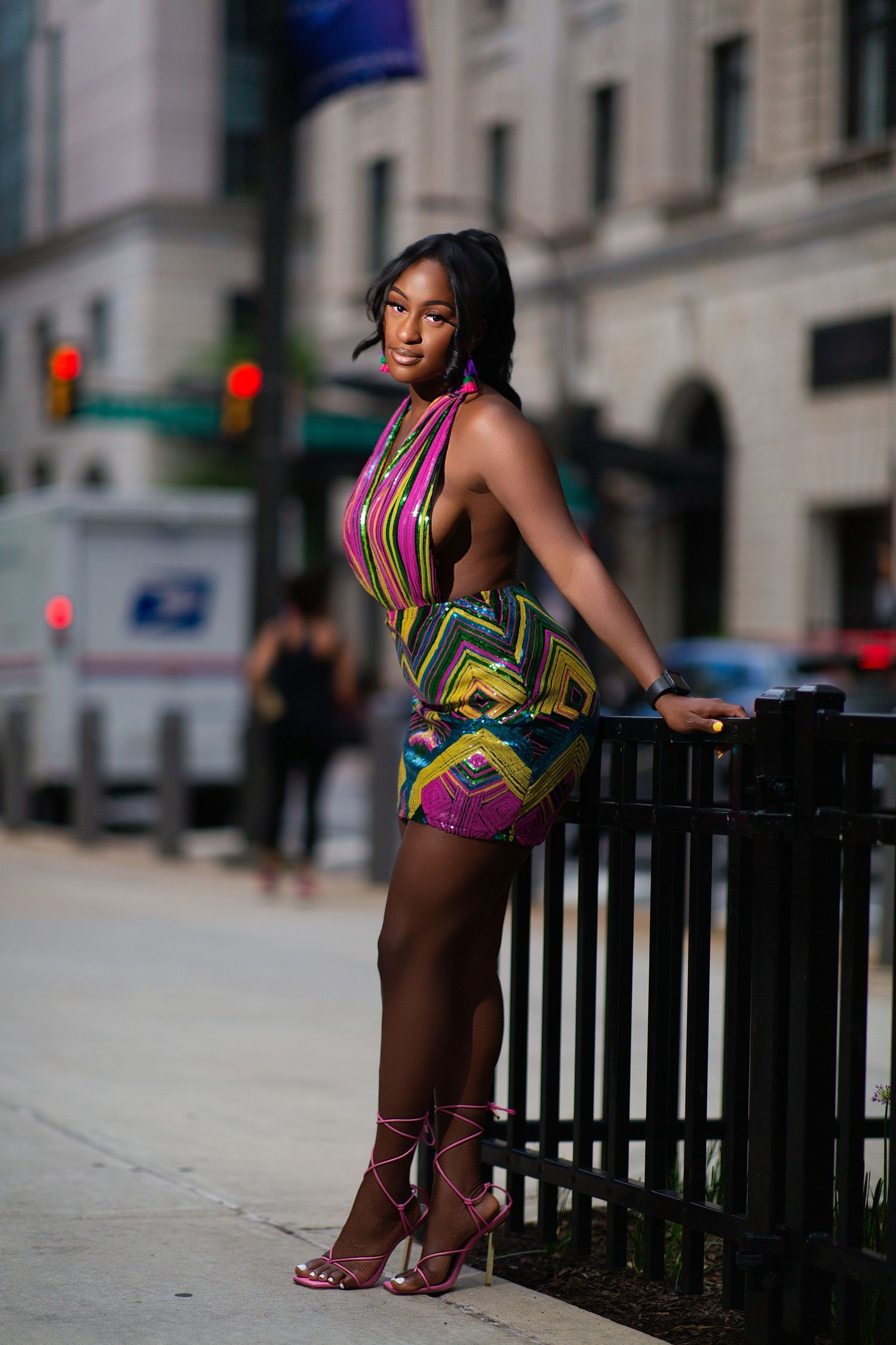 A person in a colorful printed dress poses on an urban sidewalk near a black metal fence.