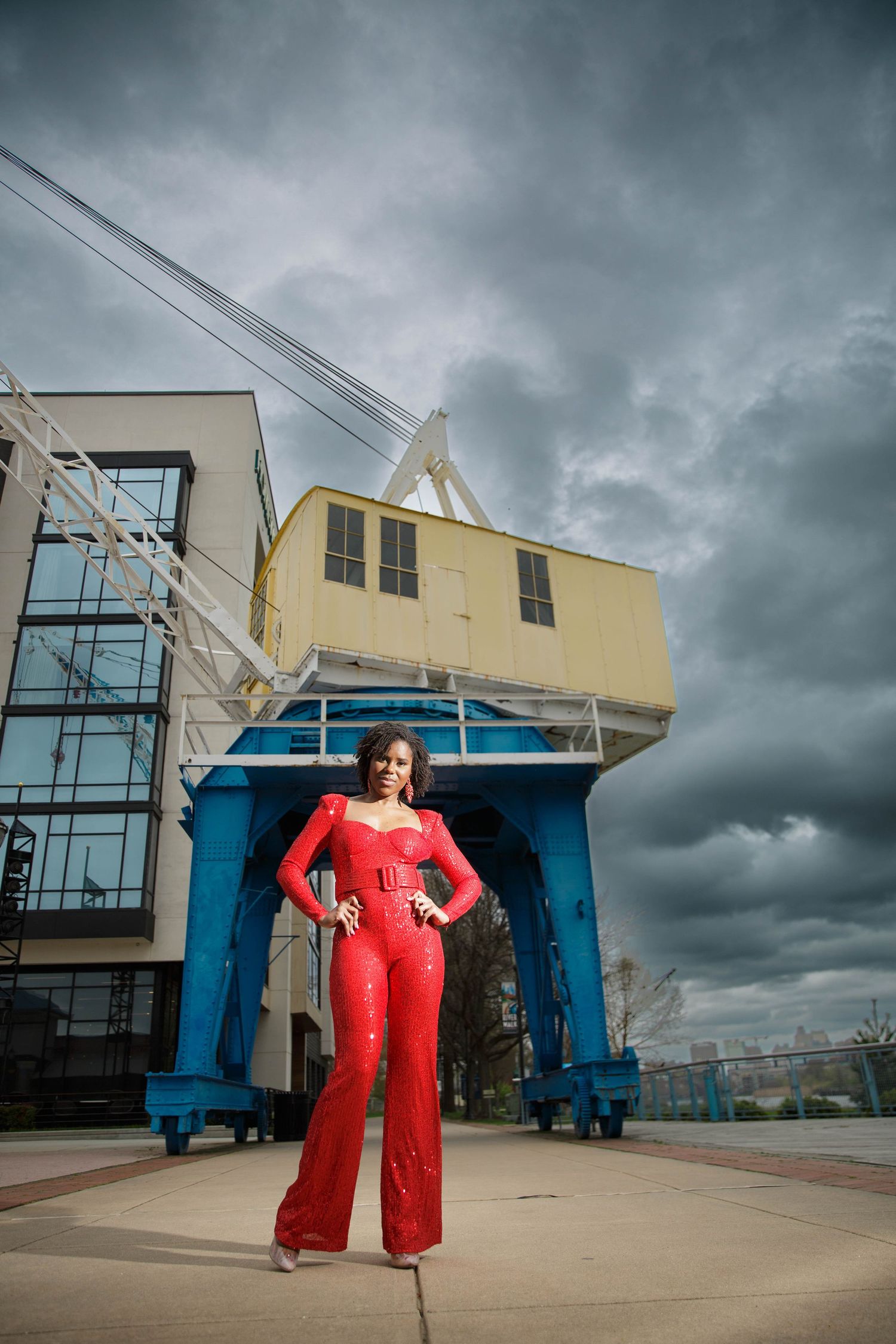 A figure in a striking red jumpsuit poses dramatically against industrial blue shipping containers under stormy skies.