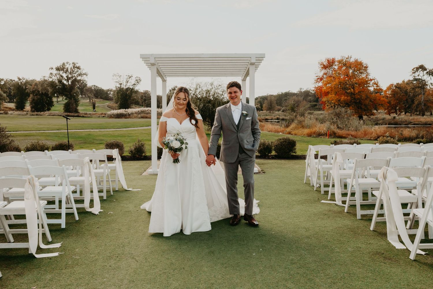 Bride and groom walk hand-in-hand across a sunlit golf course surrounded by vibrant fall foliage, with golden leaves framing the fairway
