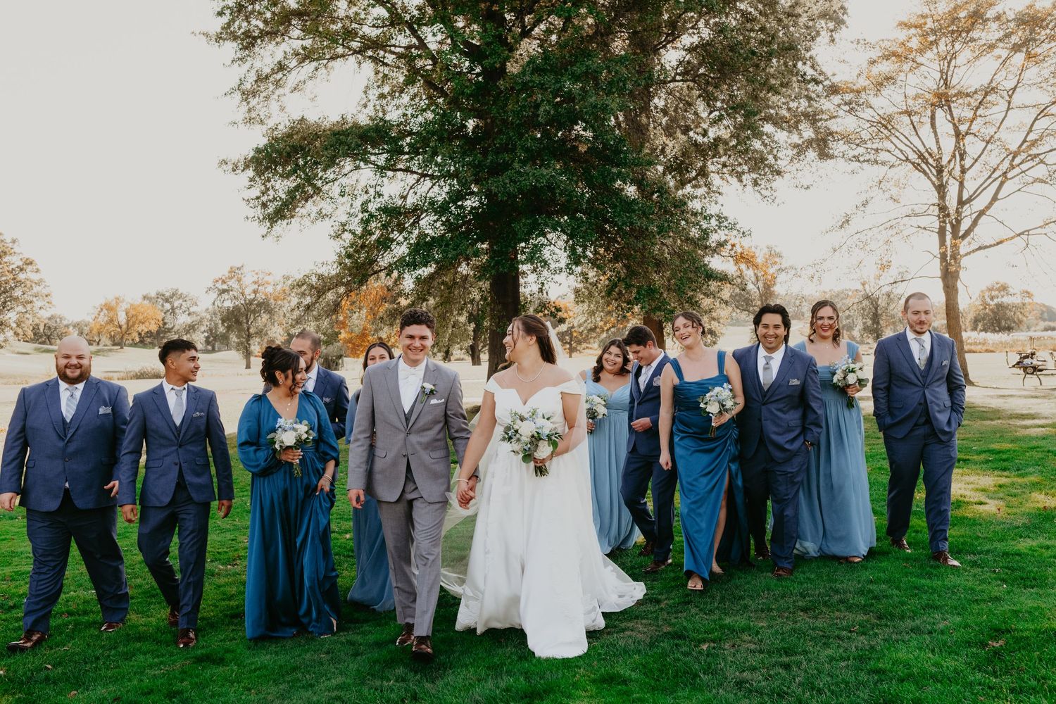 Wedding party poses on the rolling green hills of the golf course, warm fall colors stretching across the background under a soft sunset