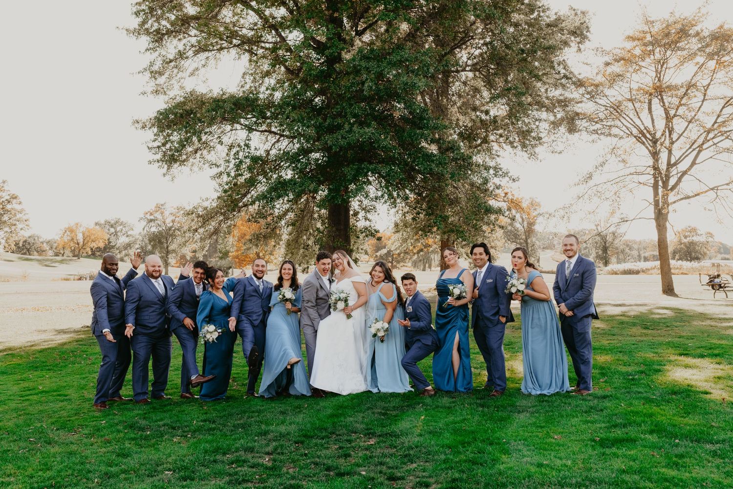 Wedding party poses on the rolling green hills of the golf course, warm fall colors stretching across the background under a soft sunset