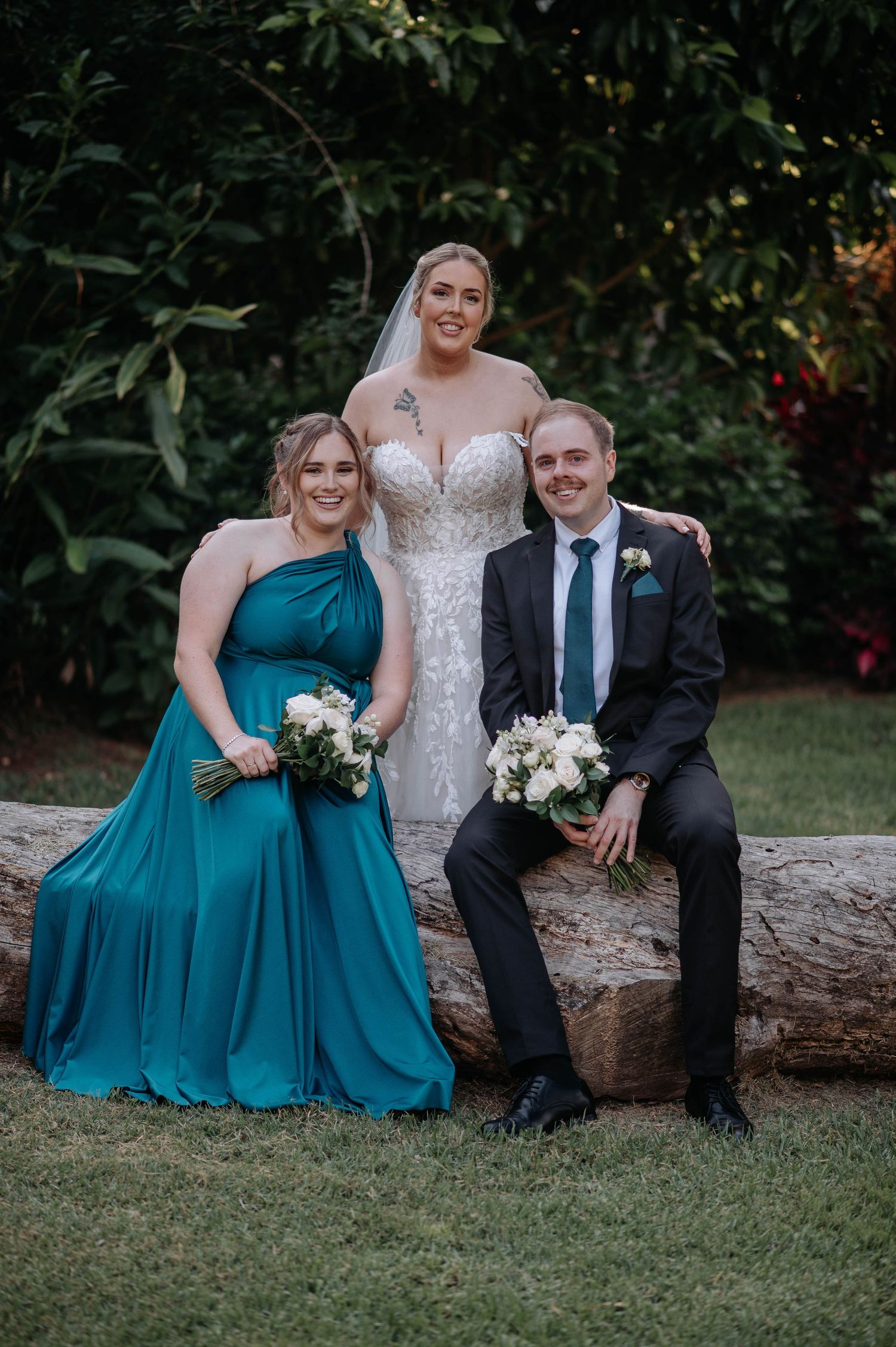 Wedding party in teal bridesmaid dresses and dark suits pose casually on fallen tree trunk in outdoor garden setting.