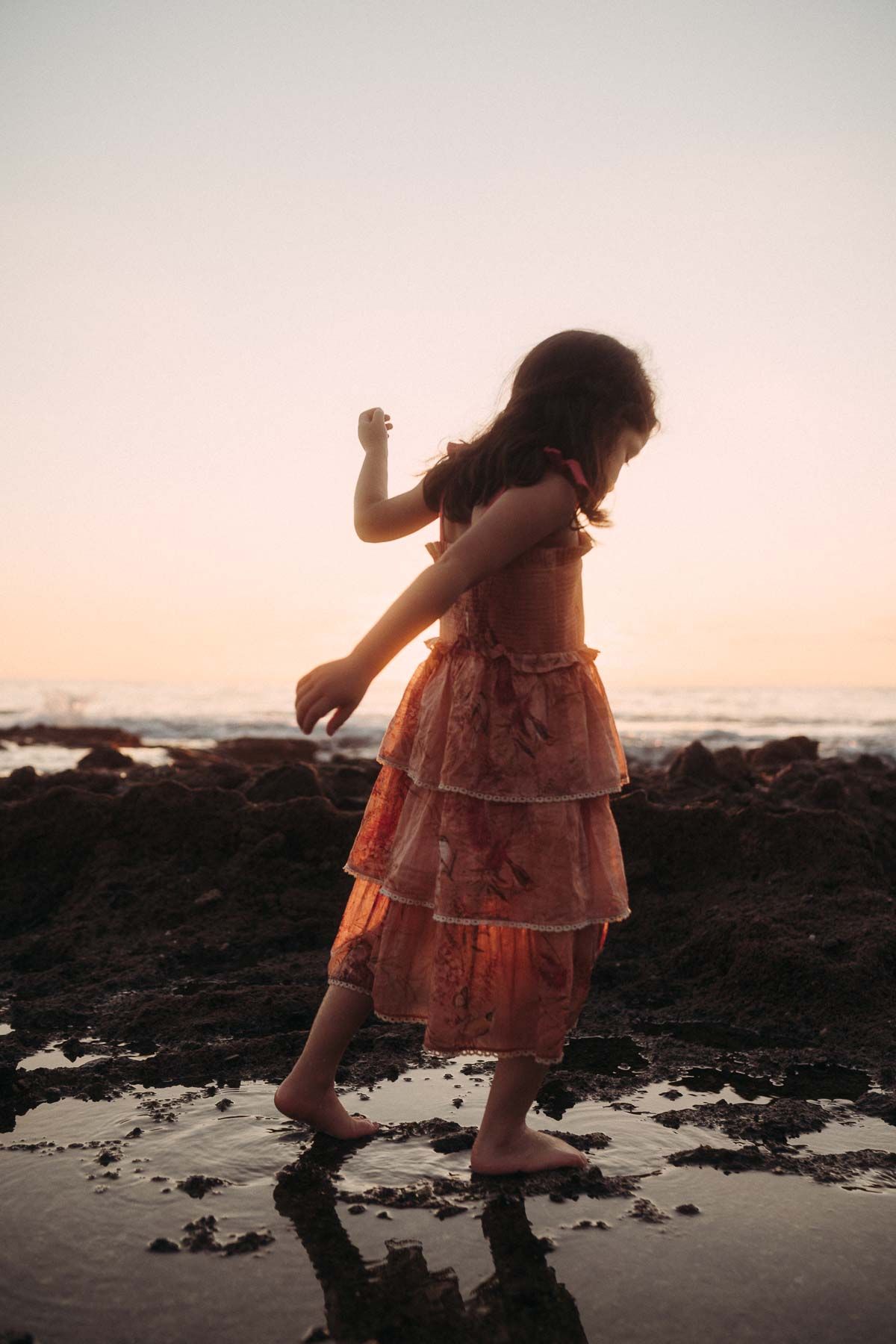 A person in a pink dress walks along a rocky beach shoreline during sunset creating a silhouetted effect.
