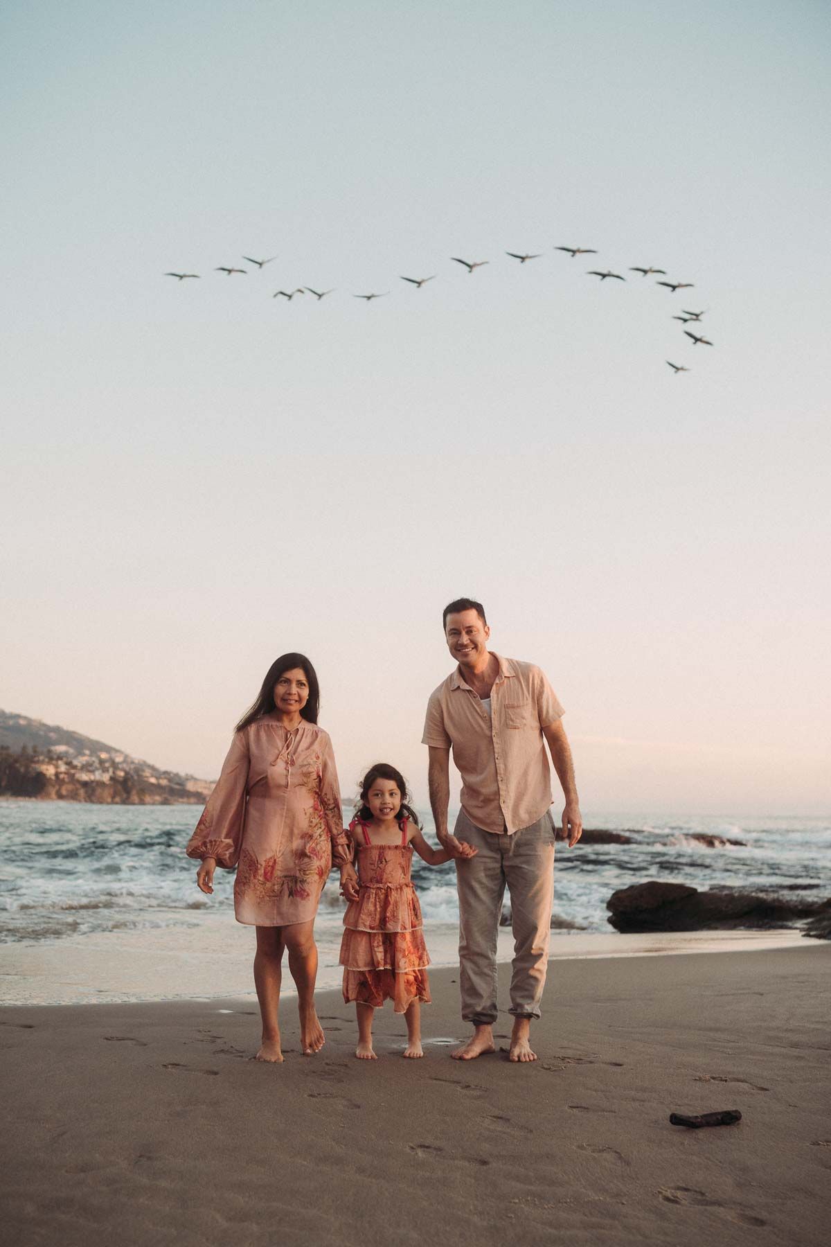 Family in coordinated outfits walking together on a scenic beach at sunset with birds flying overhead.