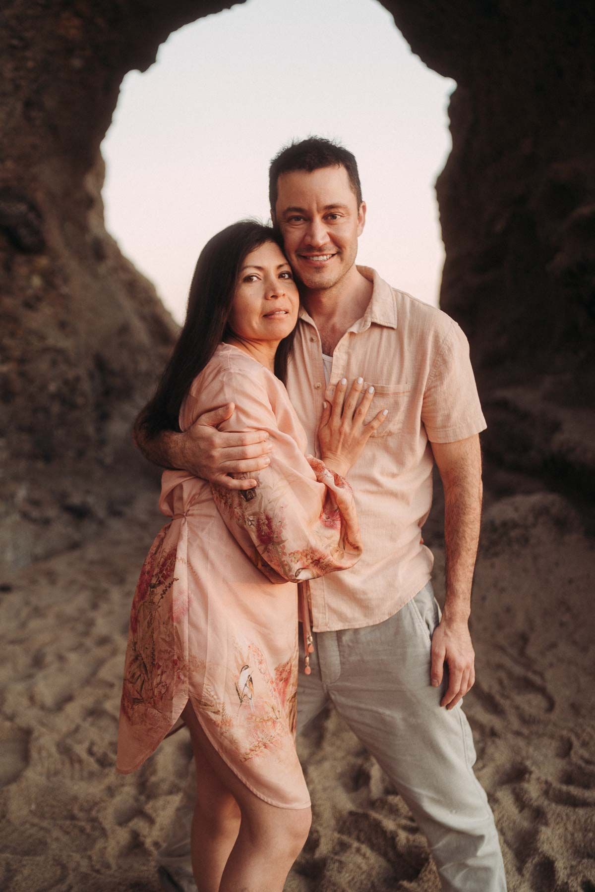 A couple in matching pink outfits embraces while posing in front of a cave opening with natural light behind them.